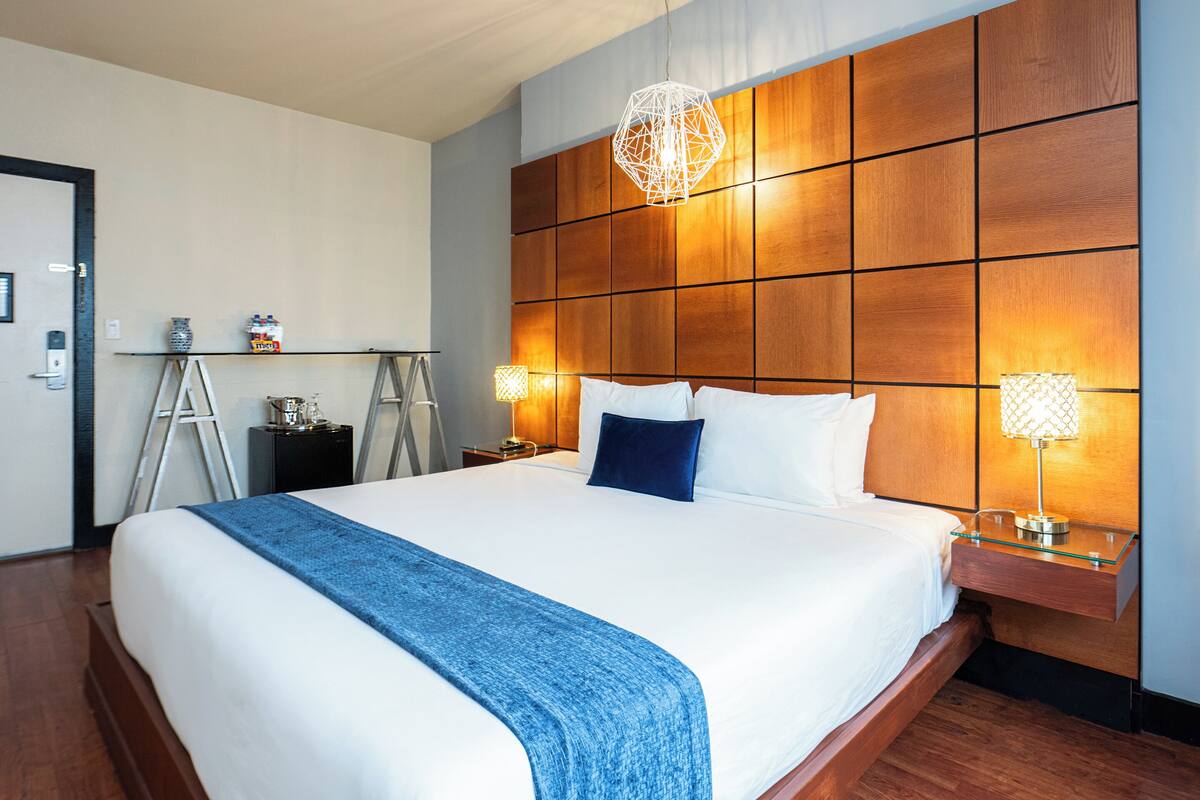 A hotel room with a large bed, wooden headboard, bedside lamps, and a blue throw blanket.