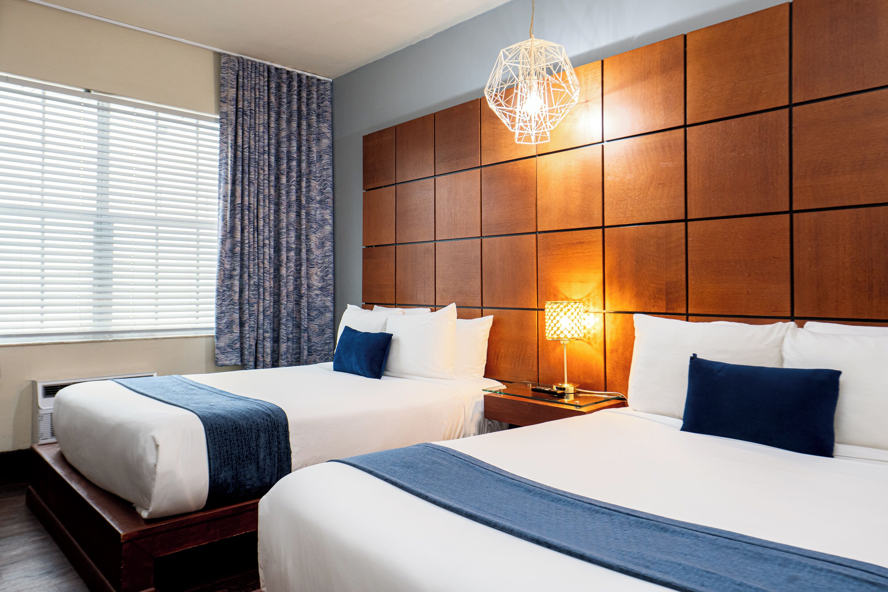 A hotel room with two beds, a wooden headboard, a bedside table with a lamp, and a window with curtains.