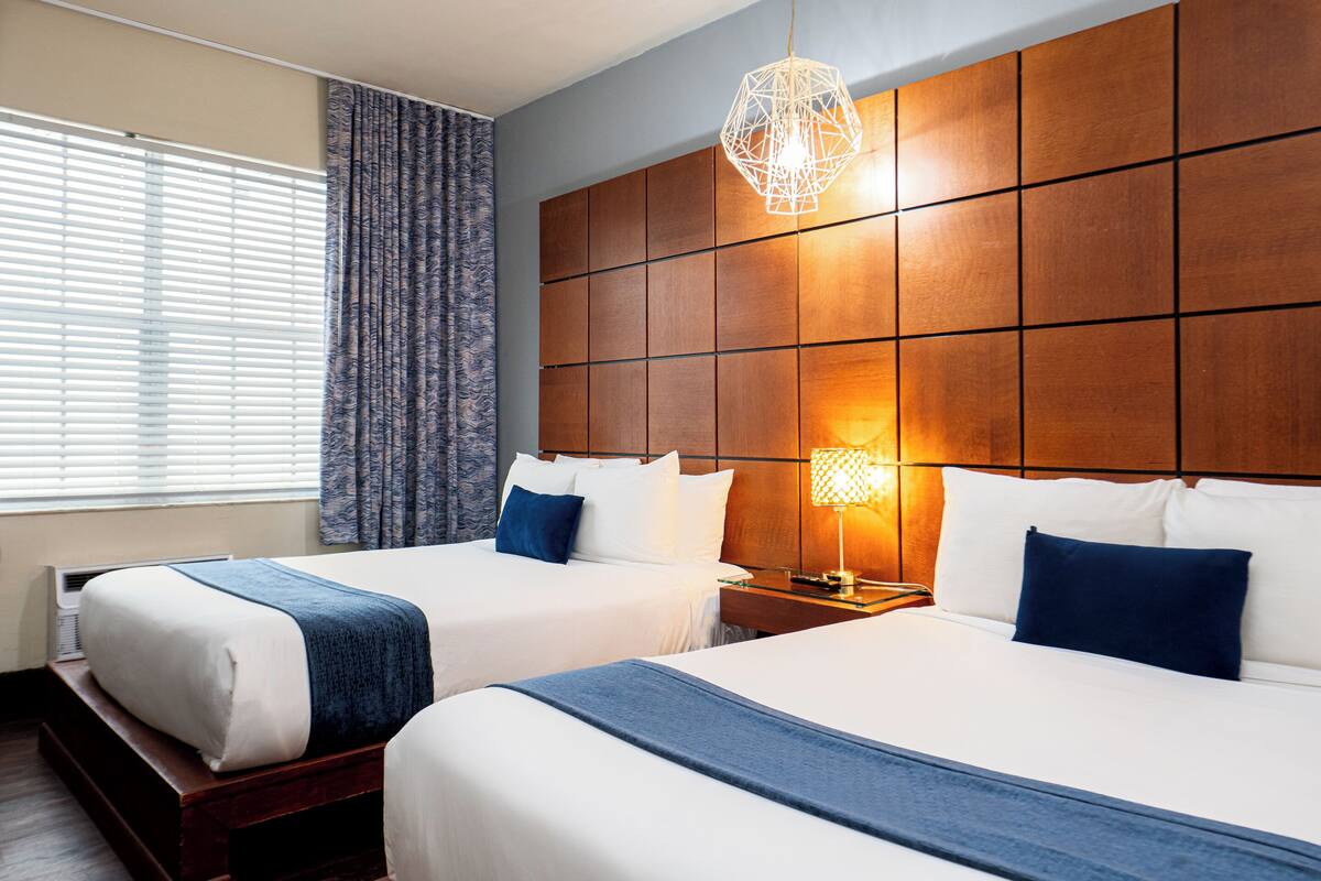 A hotel room with two beds, a wooden headboard, a bedside table with a lamp, and a window with curtains.