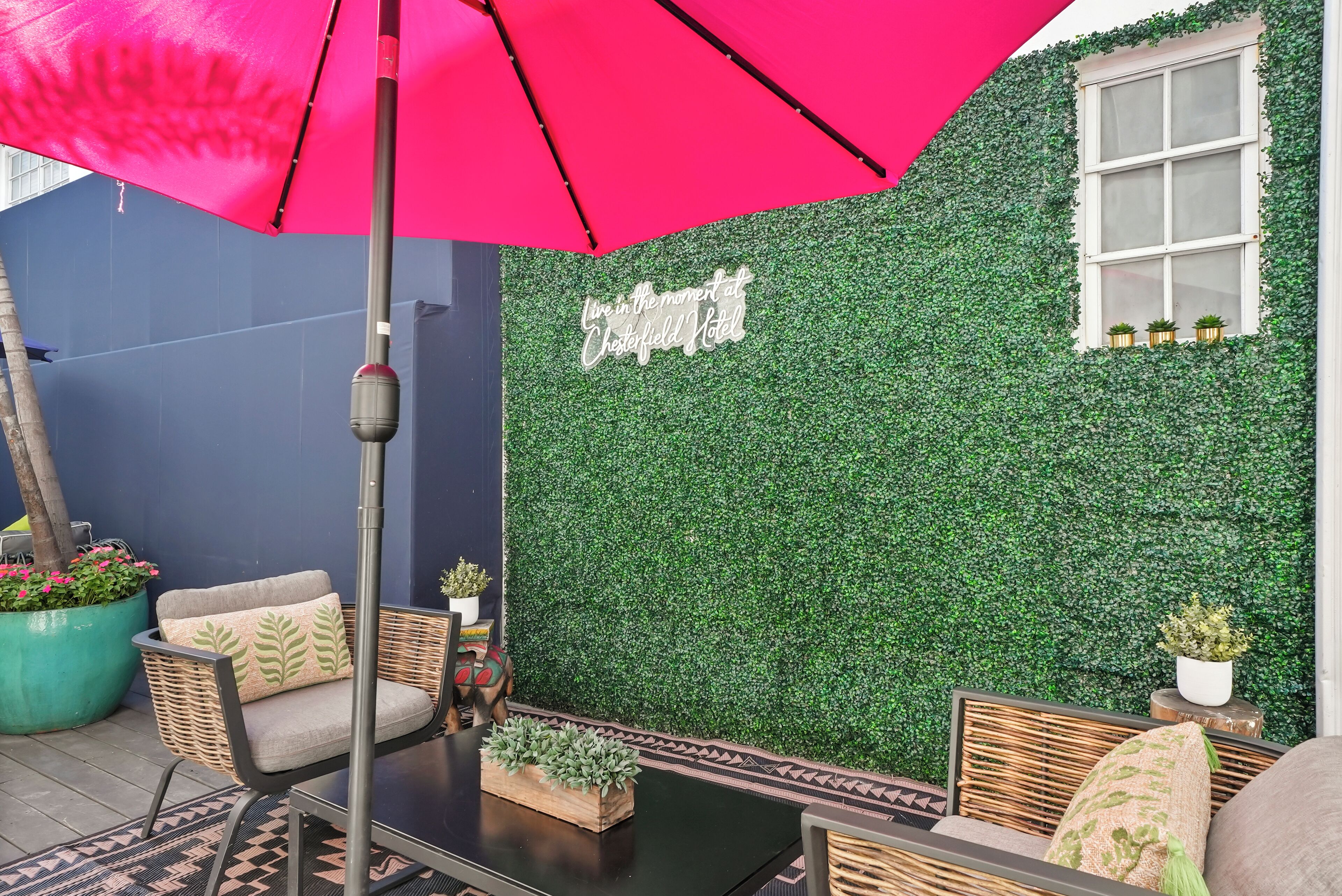 Et udendørs opholdsområde med et bord, stole og en lyserød parasol. En væg med grøn beplantning og et skilt, der siger "Live in the moment at Chesterfield Hotel" er synlig.