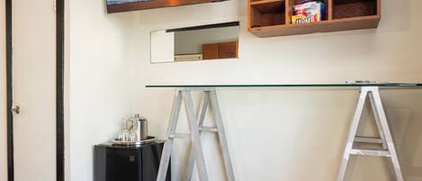 Una habitación con un escritorio de vidrio, un mueble negro pequeño, una estantería de madera con aperitivos, un espejo y una puerta.