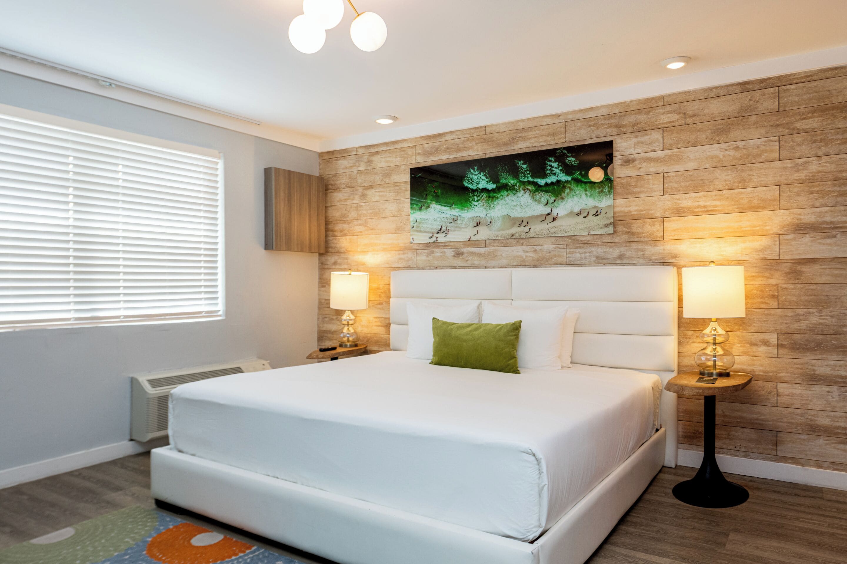 Une chambre à coucher avec un grand lit, deux tables de chevet avec des lampes, une œuvre d’art accrochée au mur et une fenêtre avec des stores.