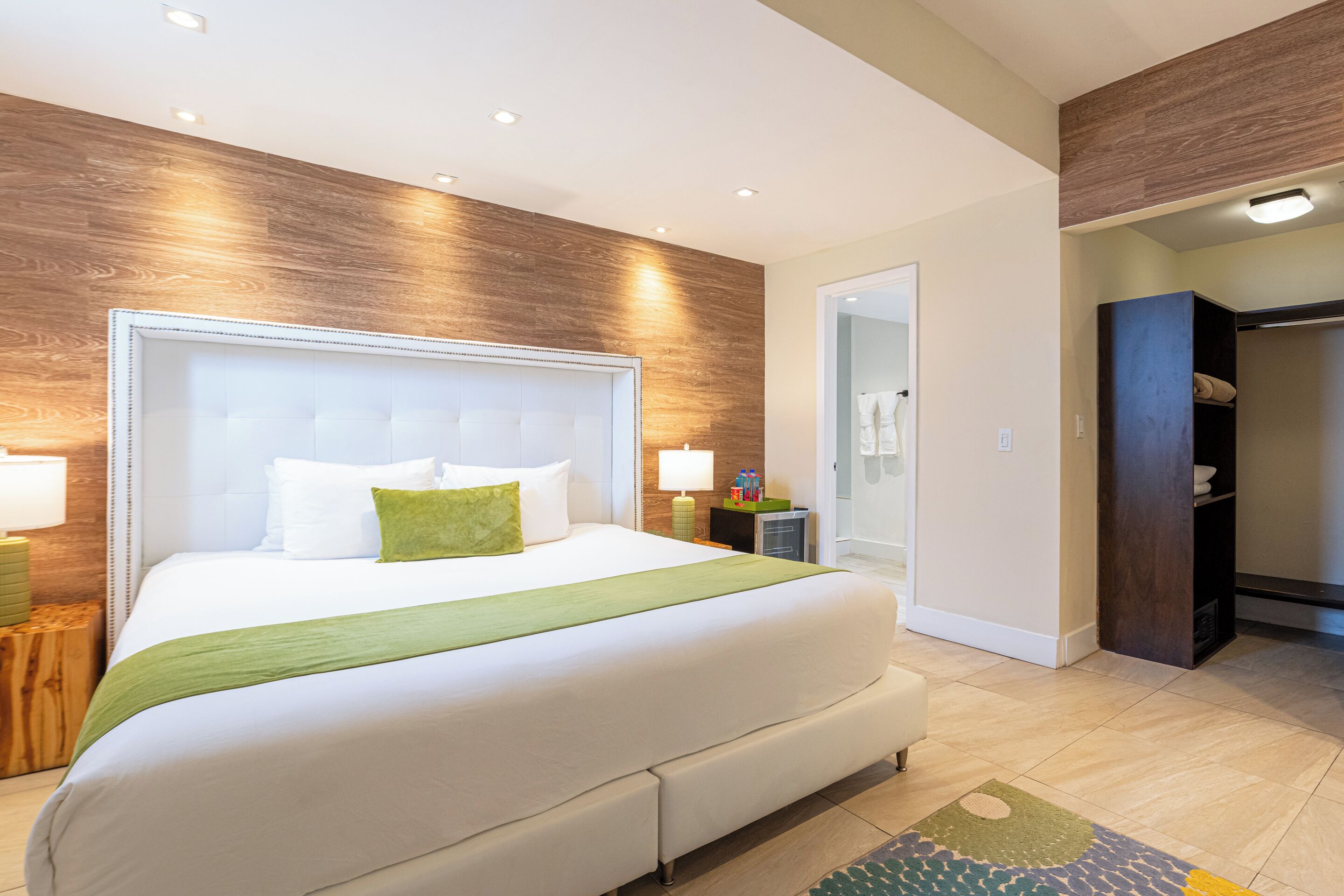 A modern hotel room with a large bed, wooden wall paneling, a walk-in closet, and a bathroom with a glass door.