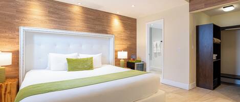 A modern hotel room with a large bed, wooden wall paneling, a walk-in closet, and a bathroom with a glass door.