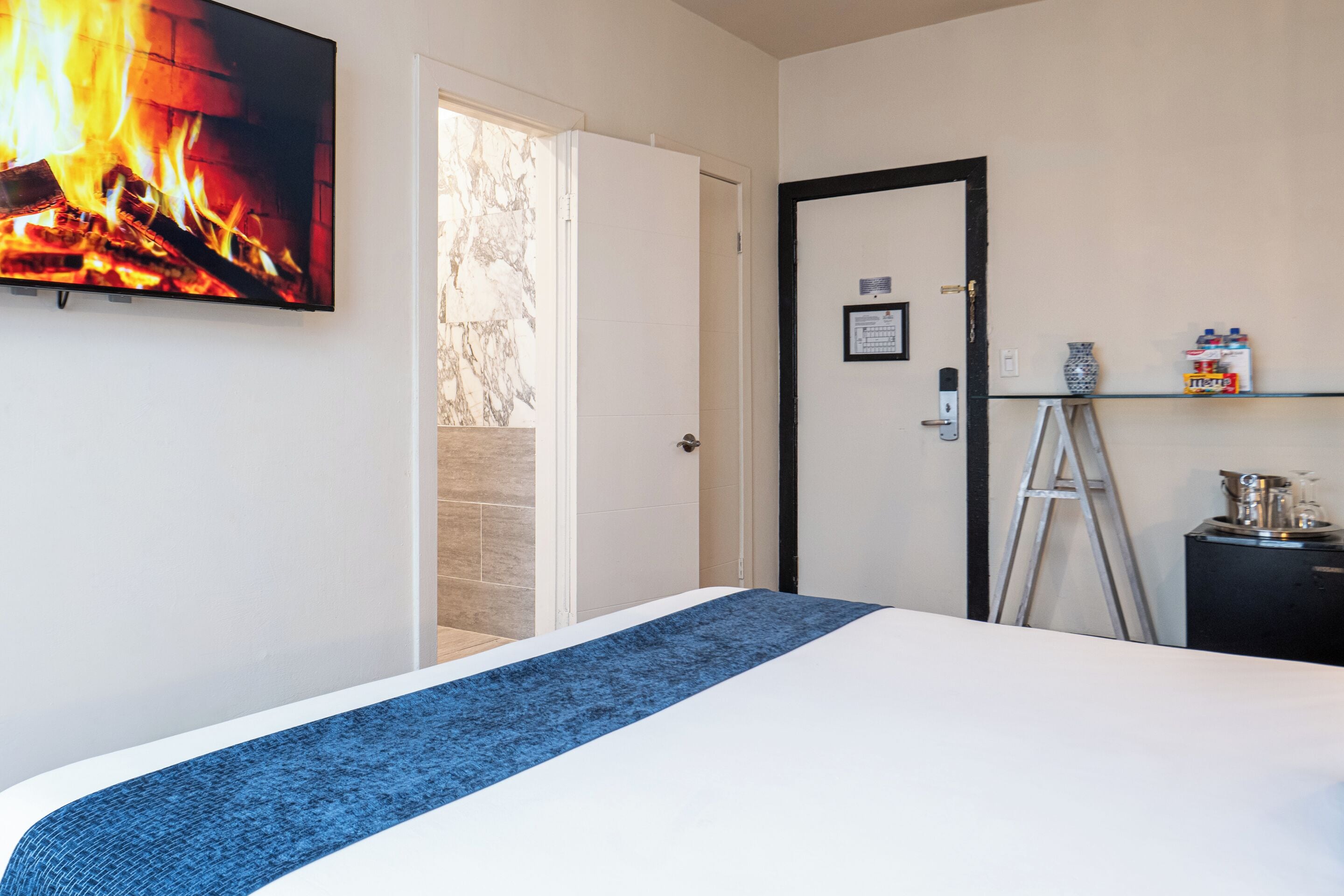 A hotel room with a bed, a television displaying a fire, a door, a ladder, and a shelf with items.