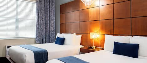 A hotel room with two beds, a wooden headboard, a nightstand with a lamp, and a window with curtains.
