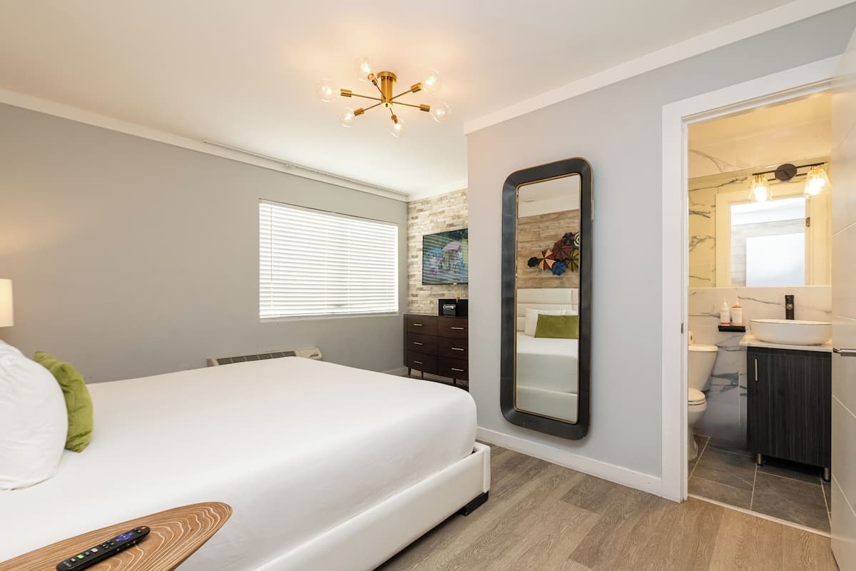 A bedroom with a bed, a bedside table, a mirror, a window, and a bathroom visible through an open door.