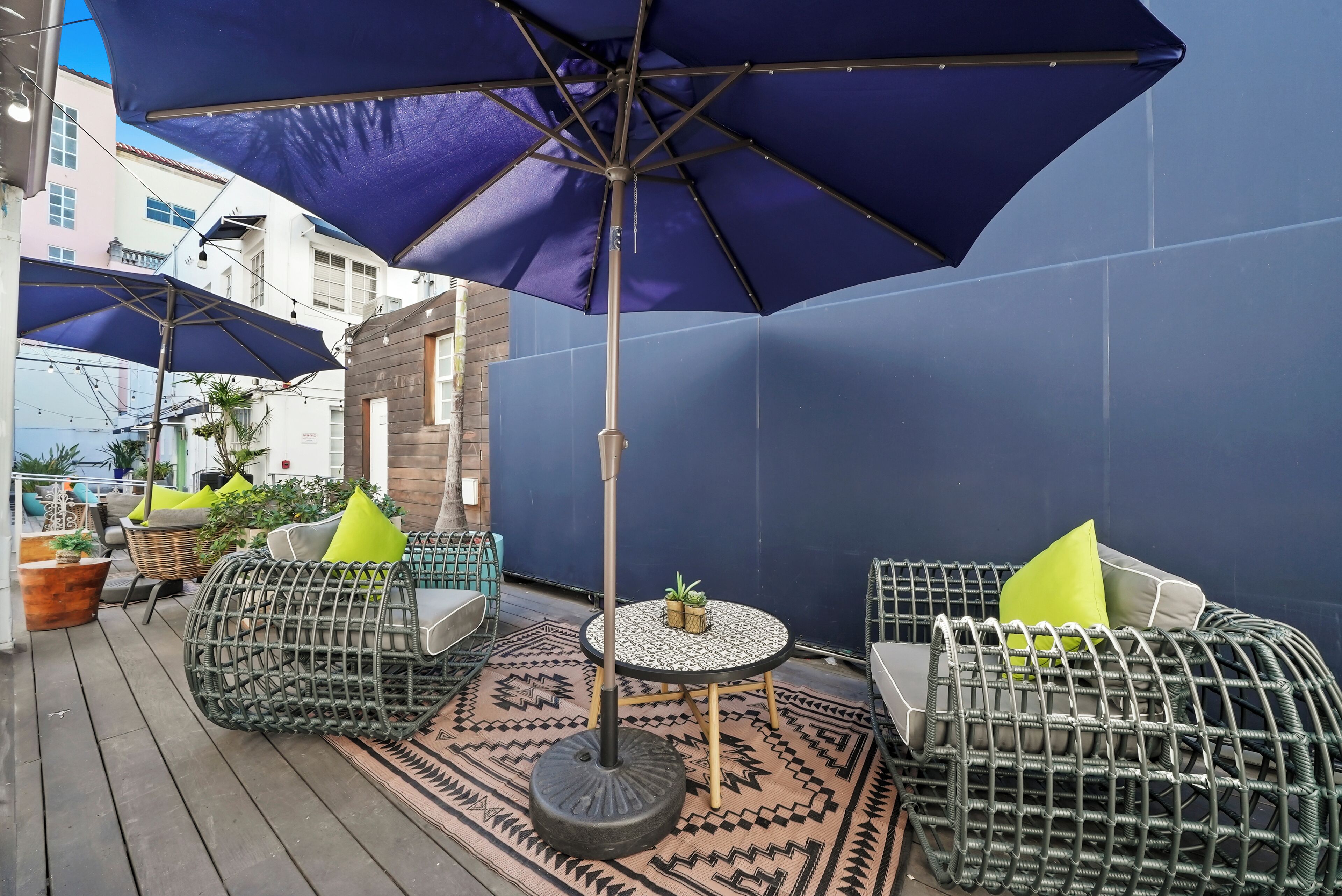 A covered outdoor seating area with wicker chairs and a round table, featuring a patterned rug and a large umbrella.