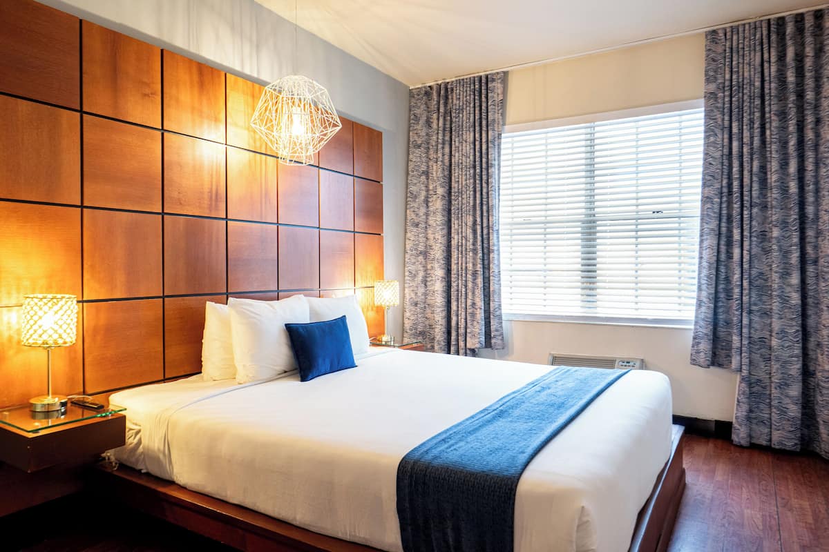 A hotel room with a large bed, wooden headboard, two bedside lamps, a window with curtains, and a wall-mounted heater.