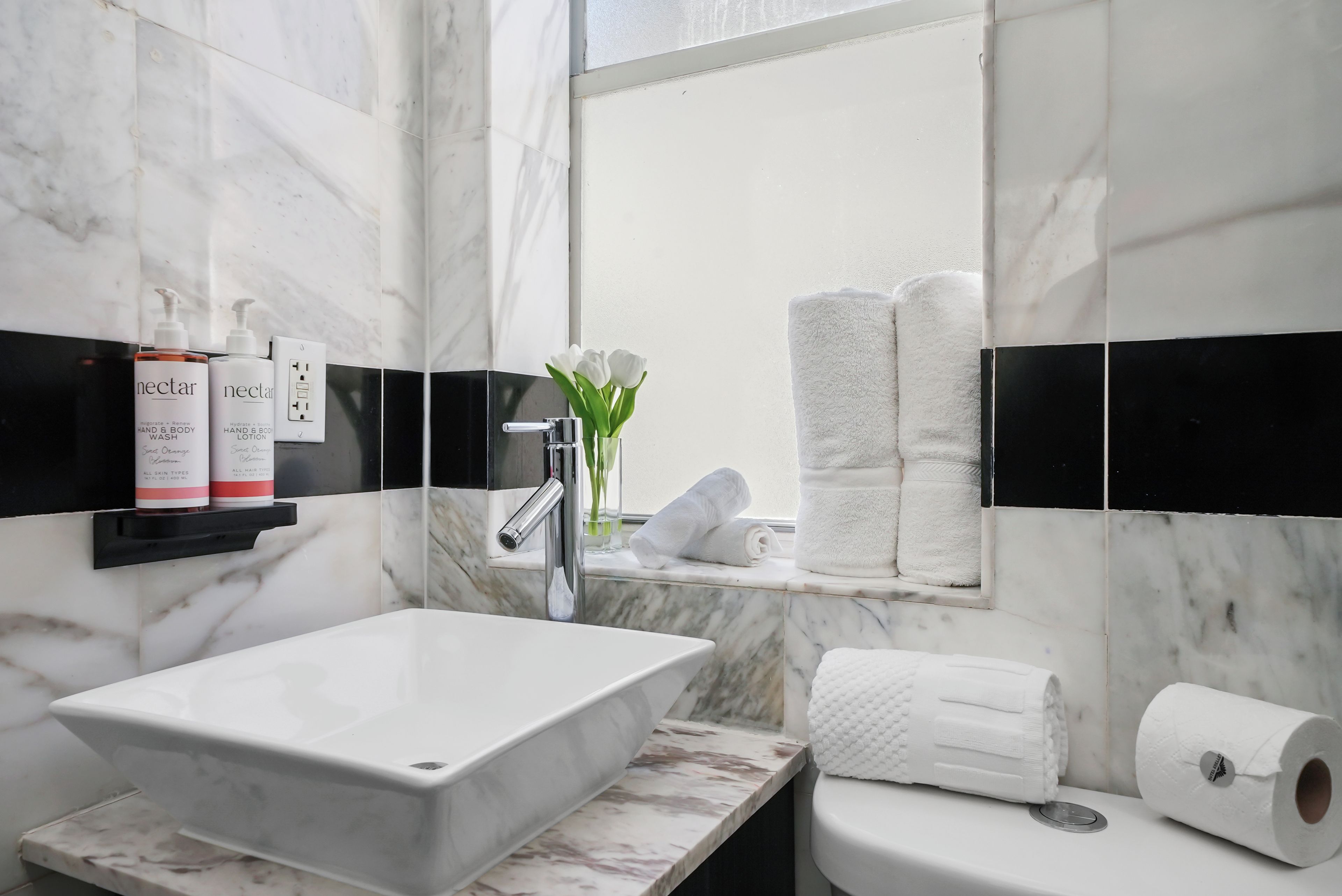 A bathroom with a marble sink, a hand soap dispenser, a hand lotion dispenser, a towel rack with towels, and a toilet paper holder.