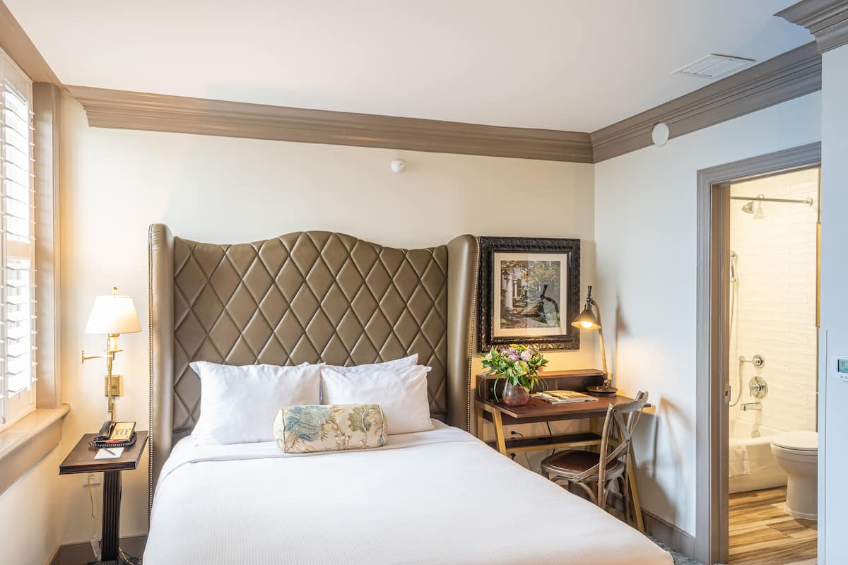 A hotel room with a large bed, a nightstand with a lamp, a chair, a desk with a computer, and a bathroom visible through an open door.