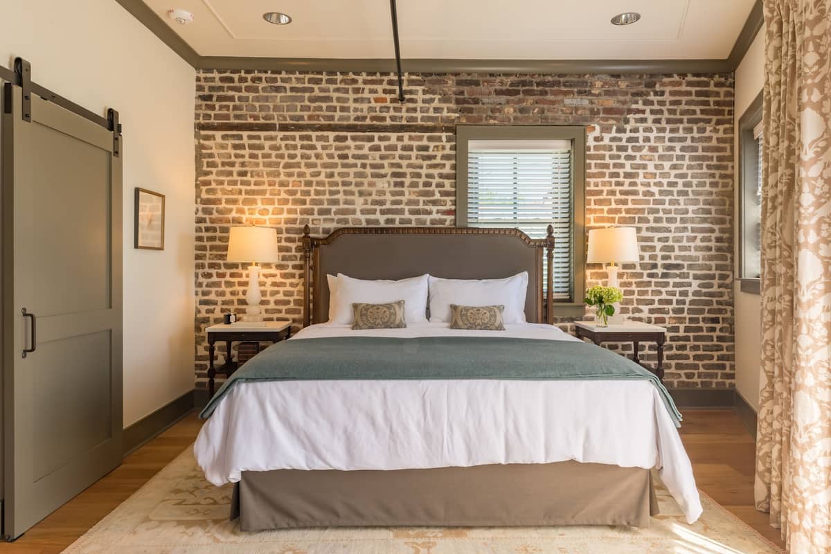 A bedroom with a brick wall, a wooden bed frame, two bedside tables with lamps, and a large window with blinds.