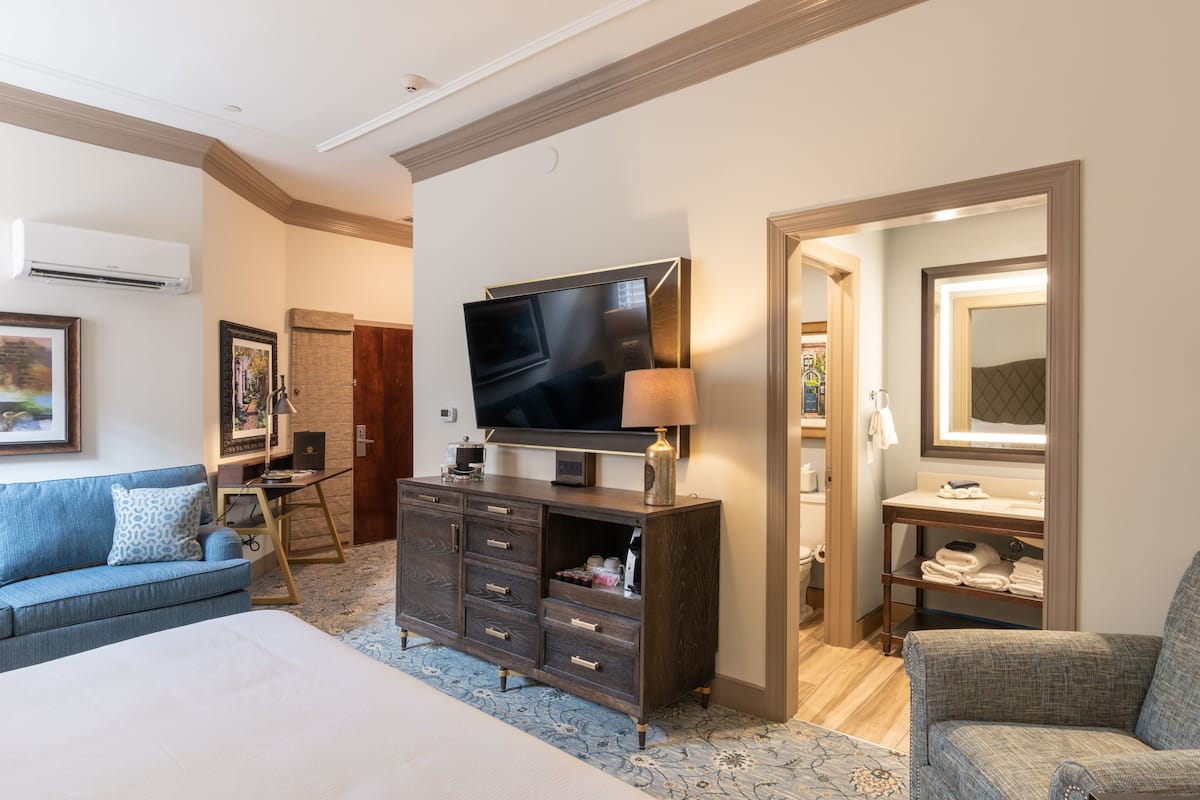 A hotel room with a large bed, a TV, a dresser, a sofa, and a bathroom visible through an open door.