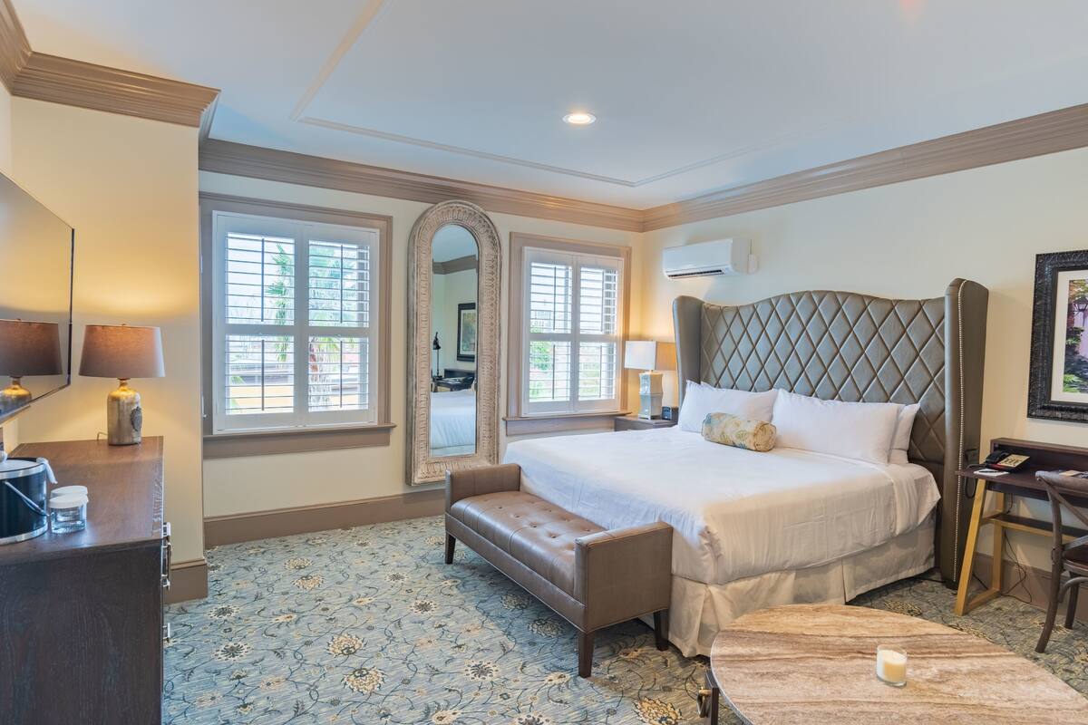 A hotel room with a large bed, a bench, a desk, and a mirror.