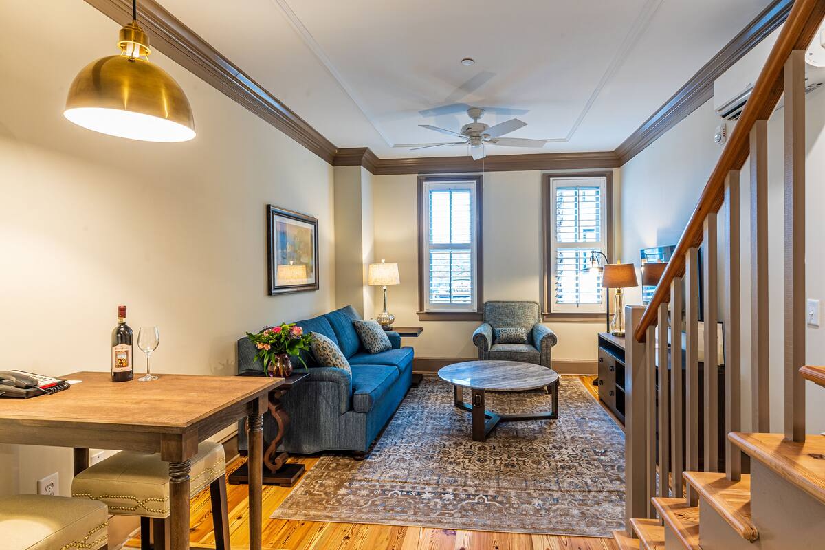 A living room with a blue sofa, a wooden coffee table, and a patterned rug. There is a ceiling fan, a window with blinds, and a staircase with wooden railings.