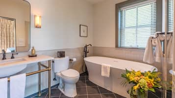 Een moderne badkamer met een wit wastafelmeubel, toilet en bad. Er is een raam met zonwering, een douchegedeelte en een vaas met bloemen.
