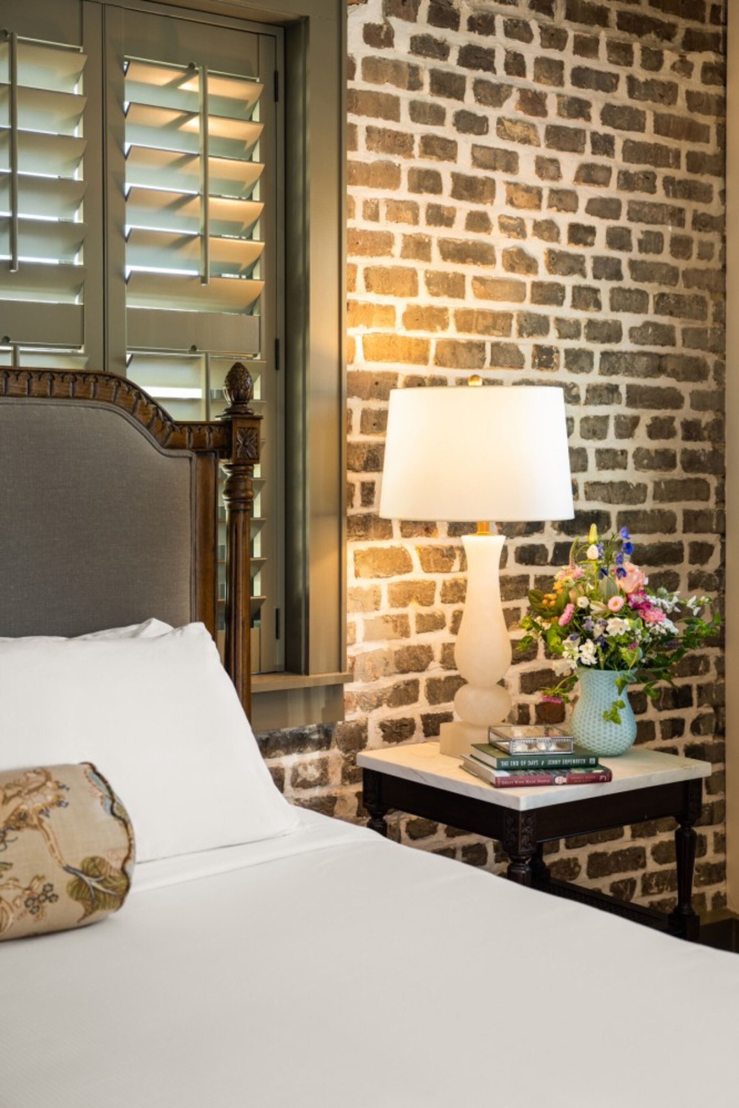 Une chambre d’hôtel avec un lit, une table de chevet avec une lampe et un vase de fleurs, et un mur en briques.