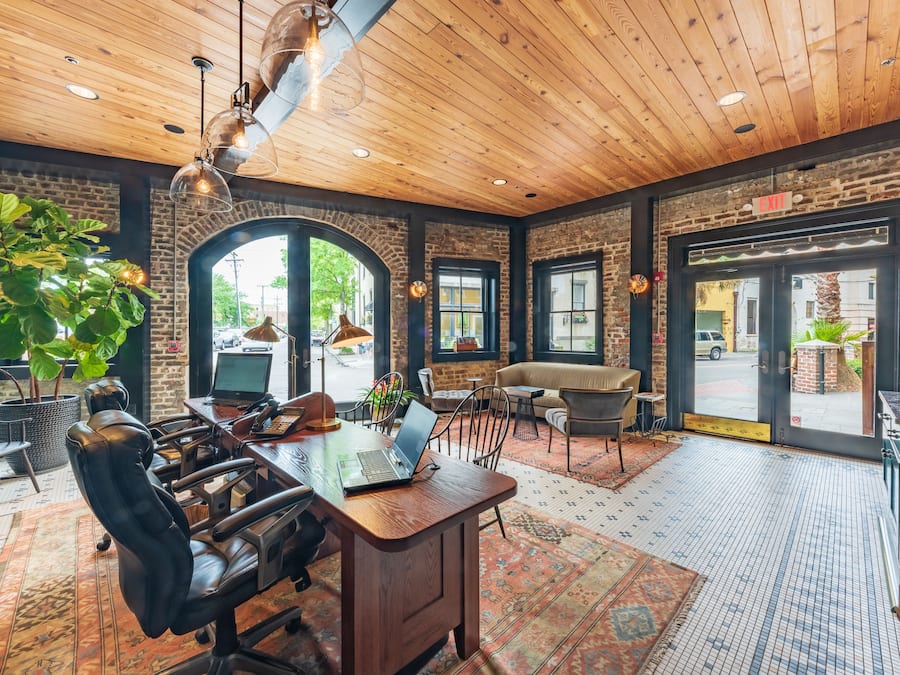 A spacious room with a wooden ceiling, a large desk with a laptop, leather chairs, and a large window with a view of the street.