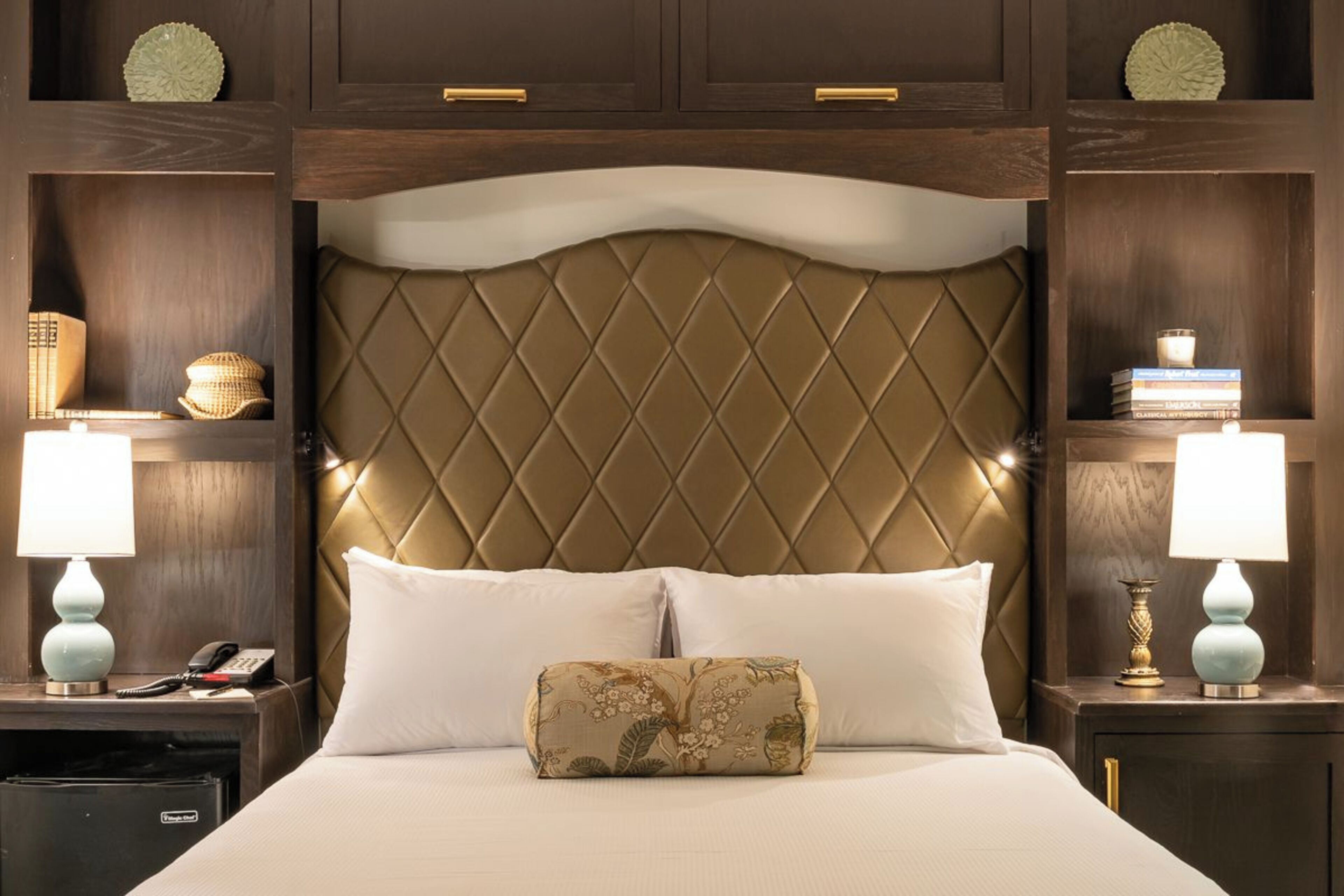 Une chambre d’hôtel avec un grand lit, deux tables de chevet, une petite table, une commode et une tête de lit ornée d’un motif en losanges.