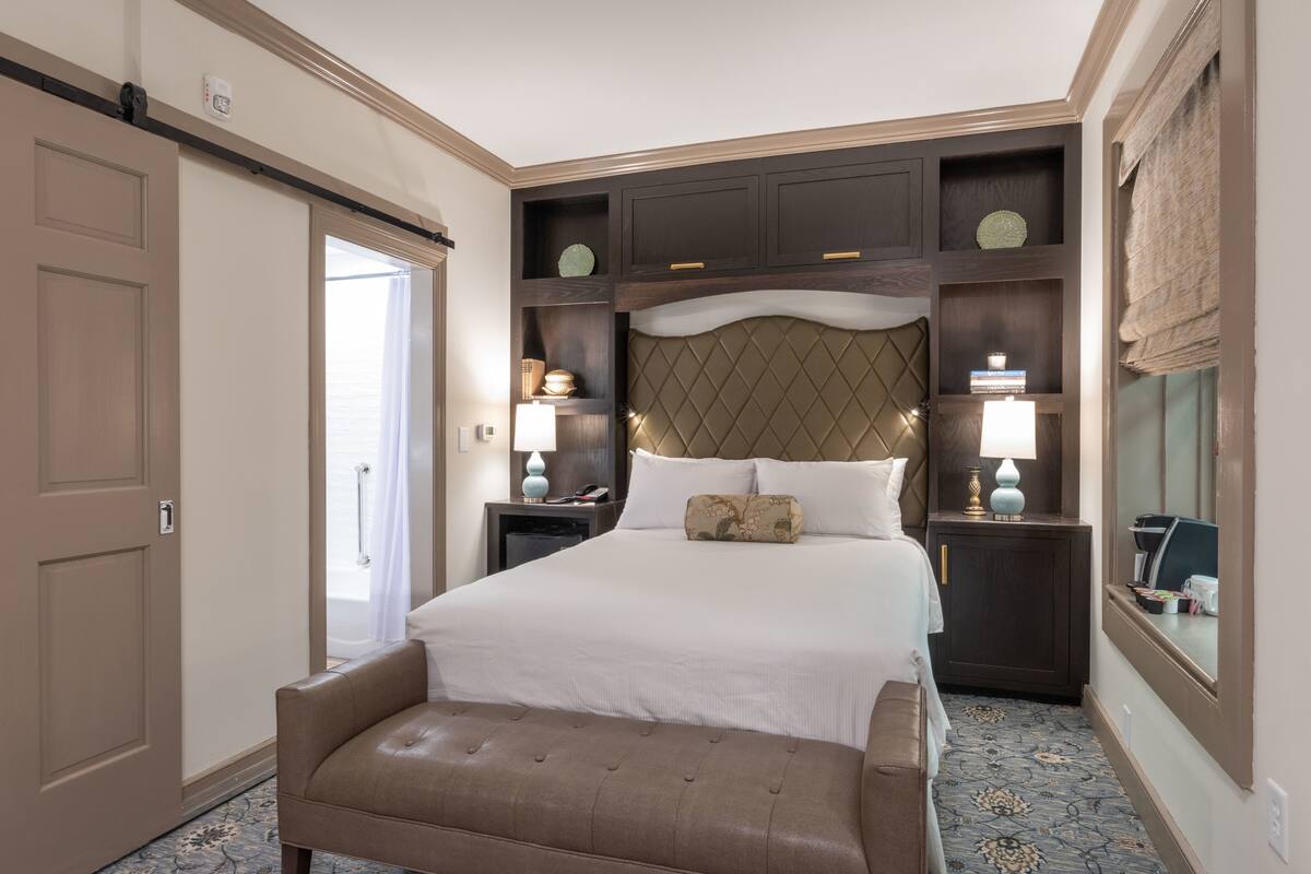 A hotel room with a large bed, a bench, a nightstand, a lamp, and a mirror.