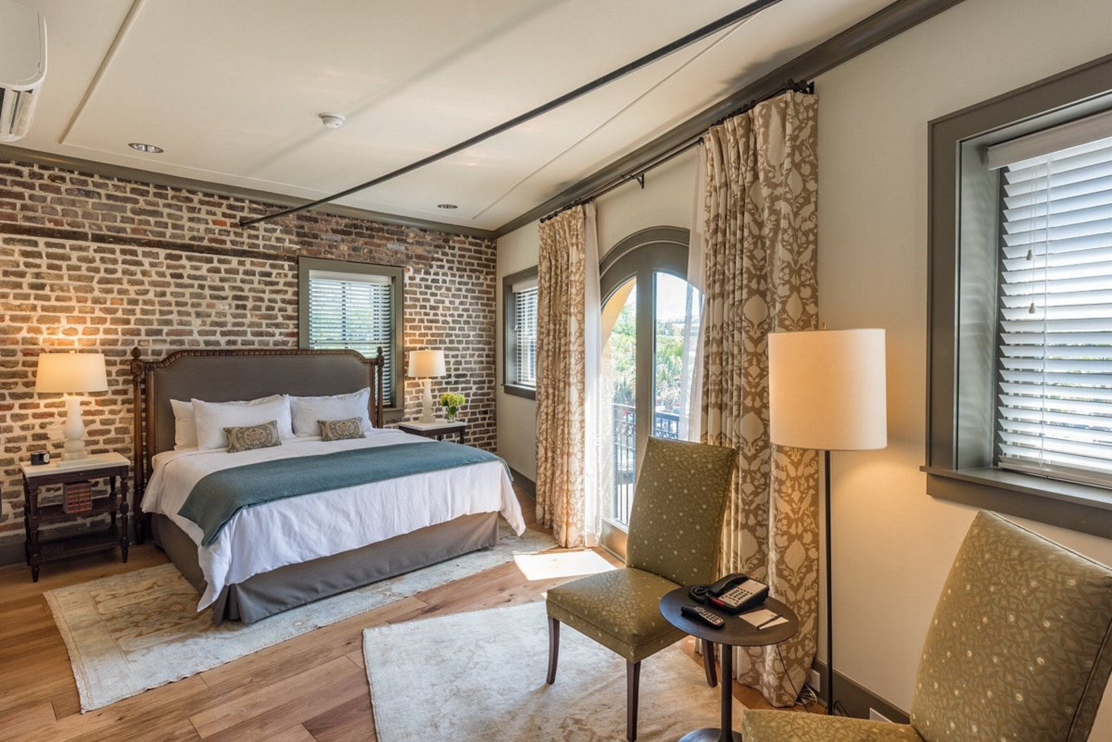 Habitación de hotel con pared de ladrillo, cama con ropa de cama blanca, una silla, una mesita con lámpara y ventana con cortinas.