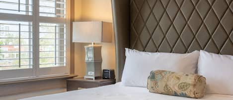 A hotel room with a large bed, a headboard, a bedside table with a lamp, and a window with blinds.