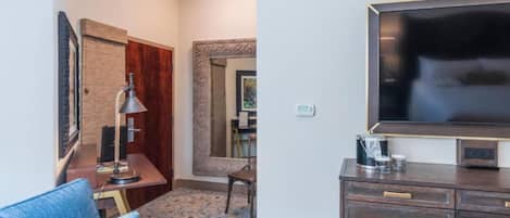 A hotel room with a large flat-screen TV, a desk with a lamp, a chair, and a coffee maker.