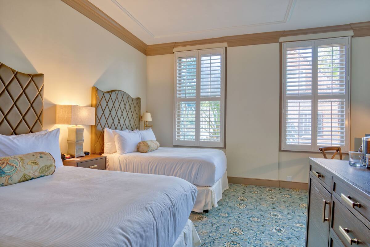 A hotel room with two beds, a dresser, a lamp, and a window with blinds.