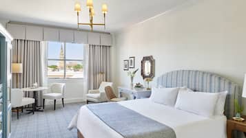 Quarto de hotel com cama grande, escrivaninha, duas cadeiras, janela com cortinas e um candelabro.
