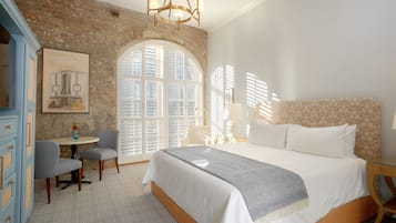 A bedroom with a large bed, a desk, and a chair. There is a window with shutters and a painting on the wall.