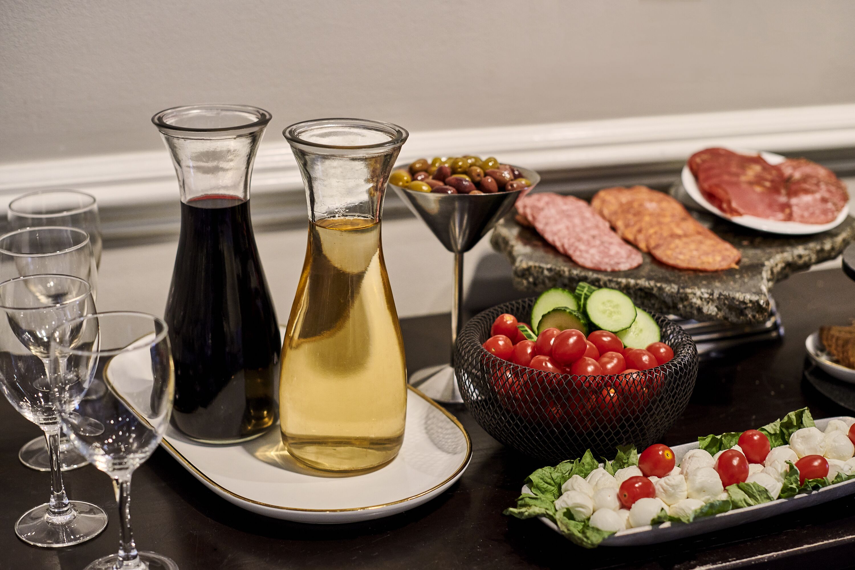 Una mesa con una botella de vino tinto, una botella de vino blanco, una copa de martini, un bol de aceitunas, un plato de salami, un plato de mozzarella y tomates, y una cesta de tomates cherry y pepinos.