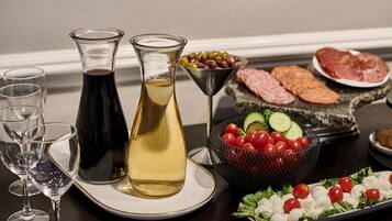 Una mesa con una botella de vino tinto, una botella de vino blanco, una copa de martini, un bol de aceitunas, un plato de salami, un plato de mozzarella y tomates, y una cesta de tomates cherry y pepinos.