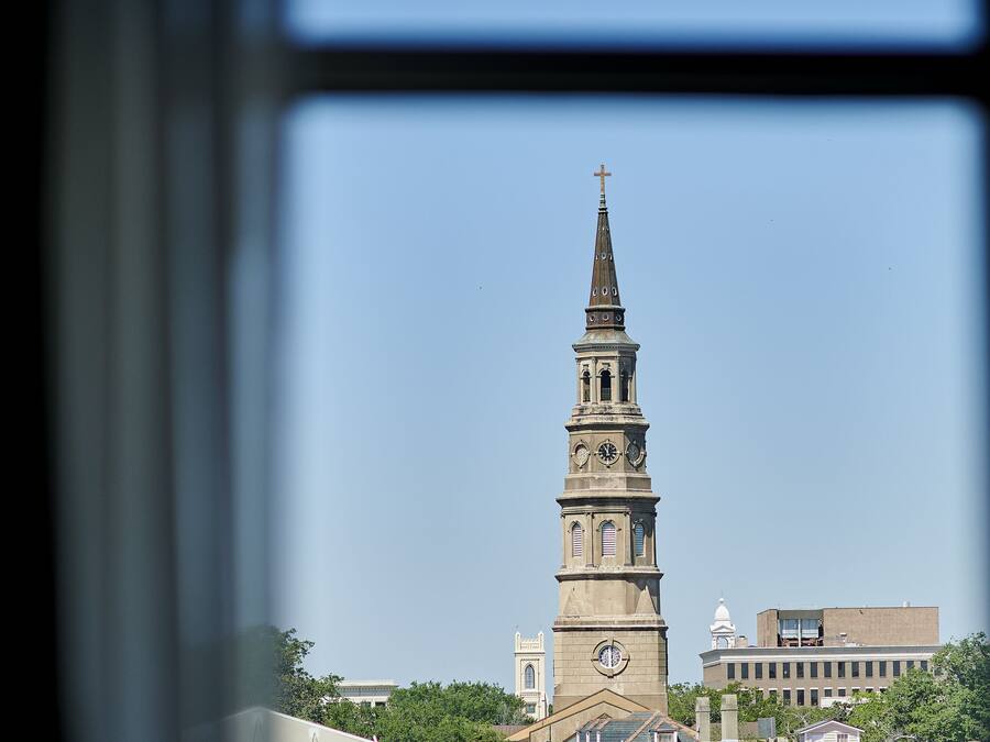 A church tower with a clock and a cross, surrounded by buildings and trees.