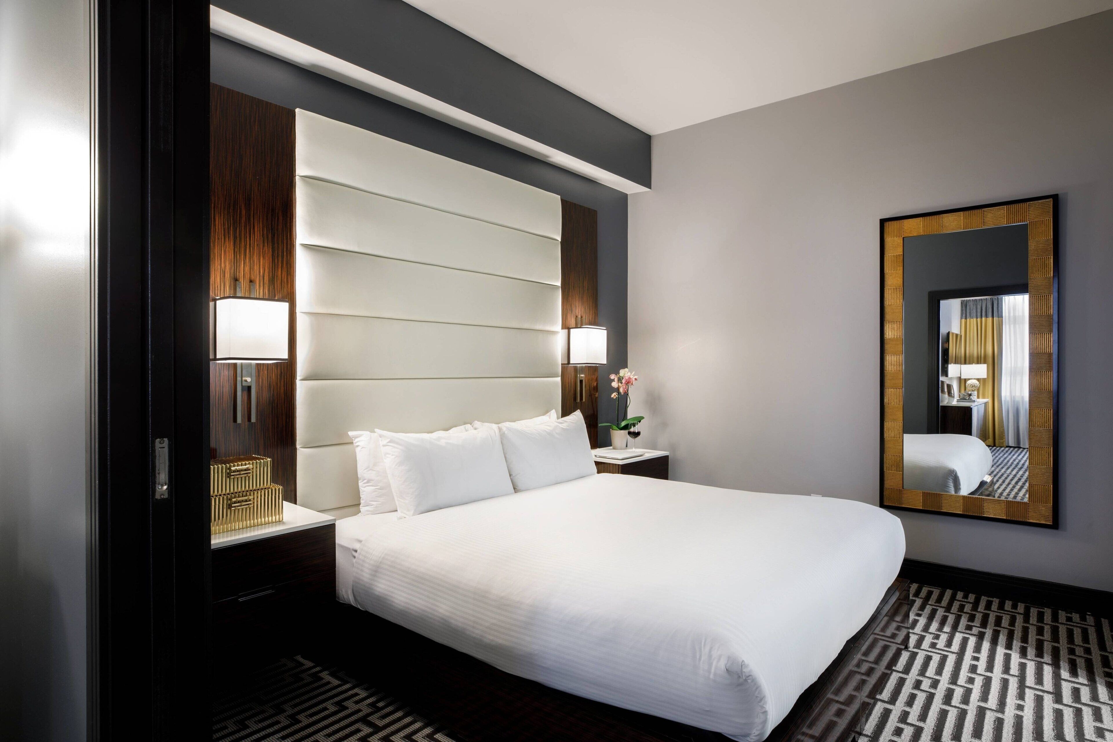 A hotel room with a large bed, a nightstand with a lamp, a mirror, and a patterned rug.
