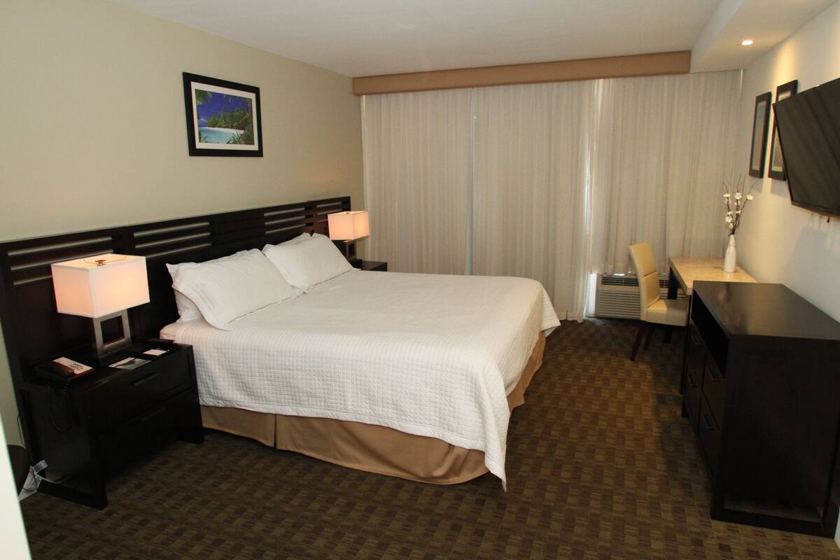 Ein Hotelzimmer mit einem großen Bett, zwei Nachttischen mit Lampen, einem Schreibtisch mit Stuhl und einem großen Fenster mit Vorhängen.