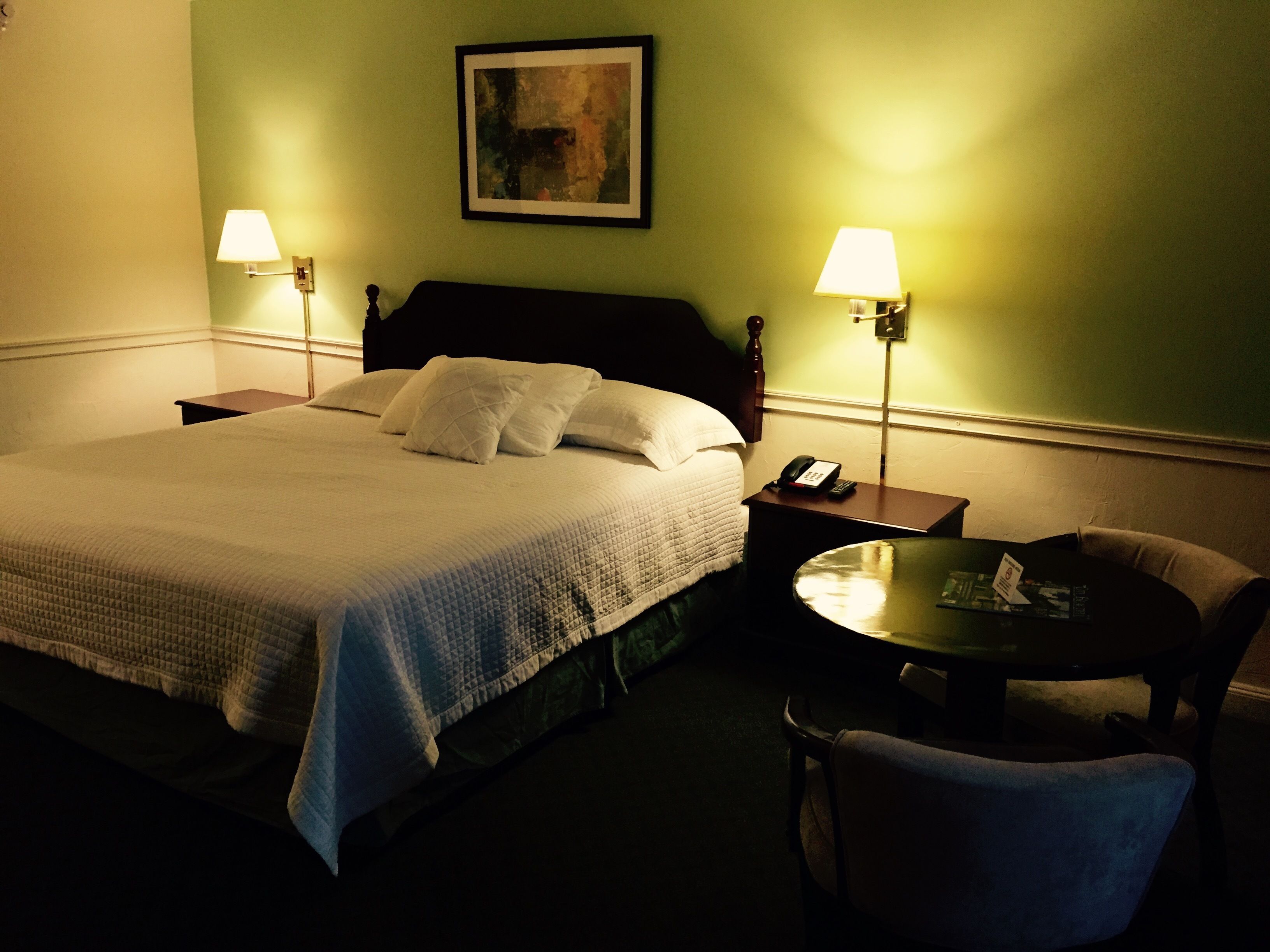 A hotel room with a bed, two bedside lamps, a chair, a small table, and a painting on the wall.