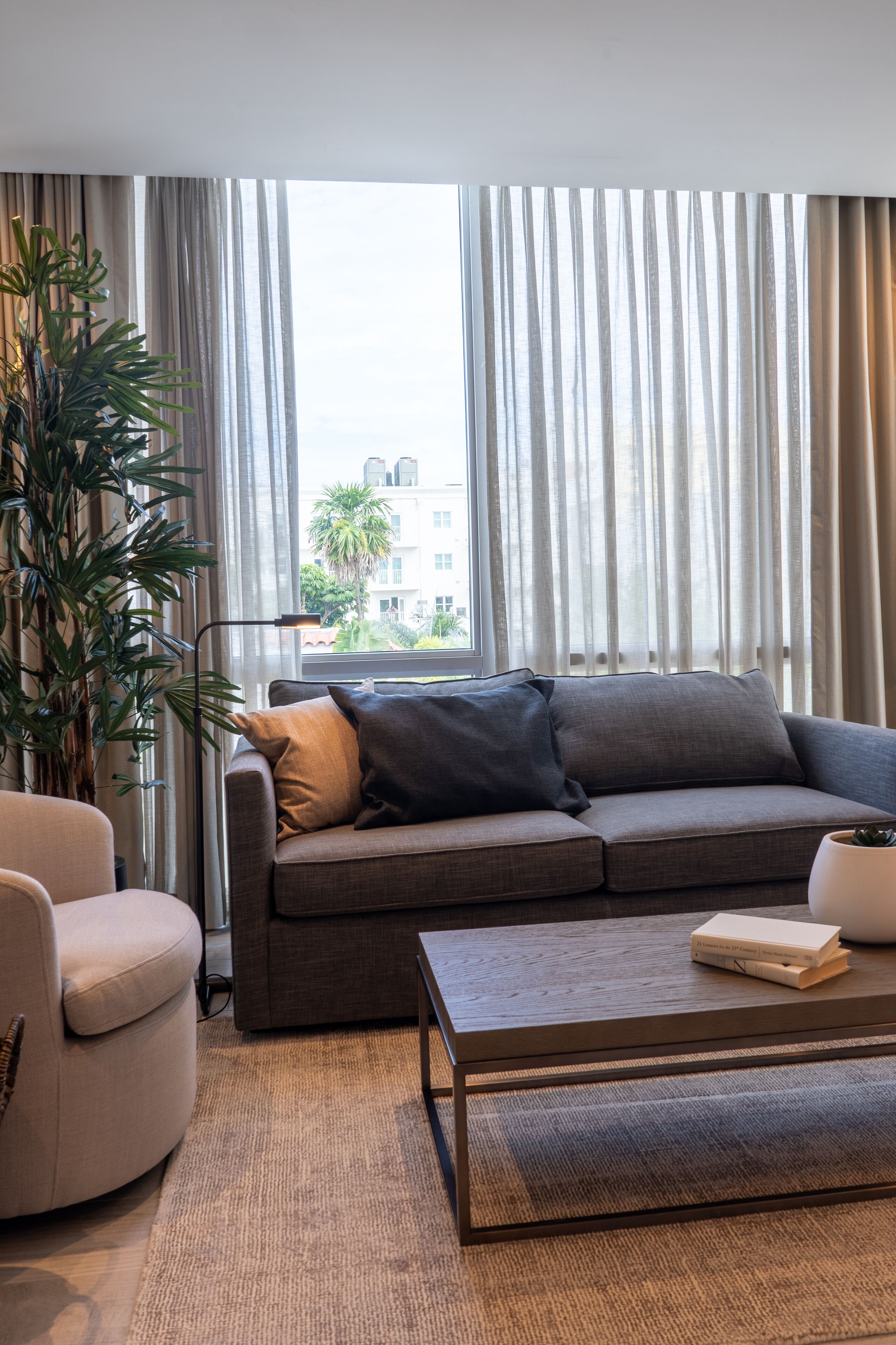 Un salon moderne avec un canapé gris, un fauteuil blanc, une table basse et une grande plante.