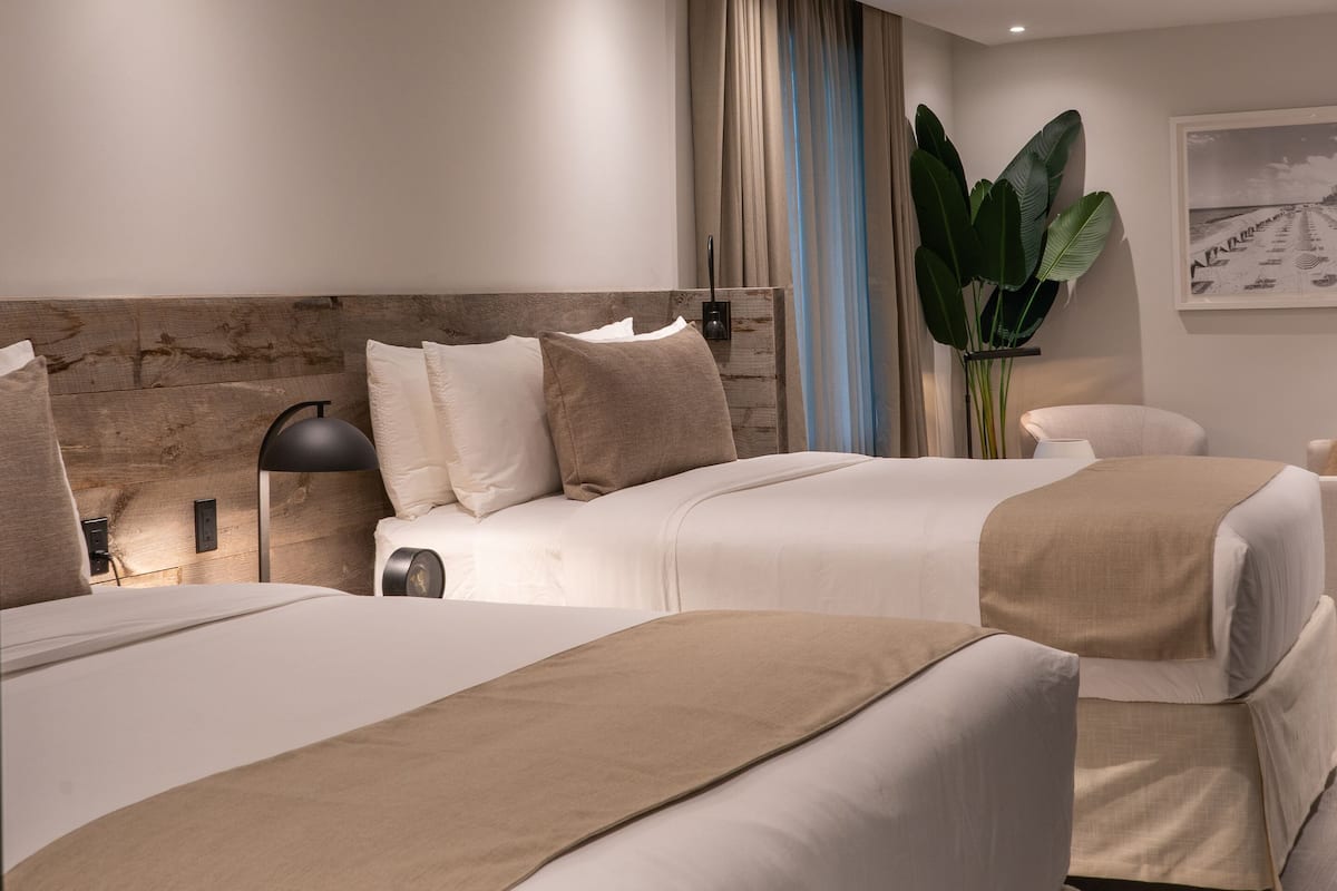 A hotel room with two beds, a wooden headboard, a large plant, and a framed picture on the wall.