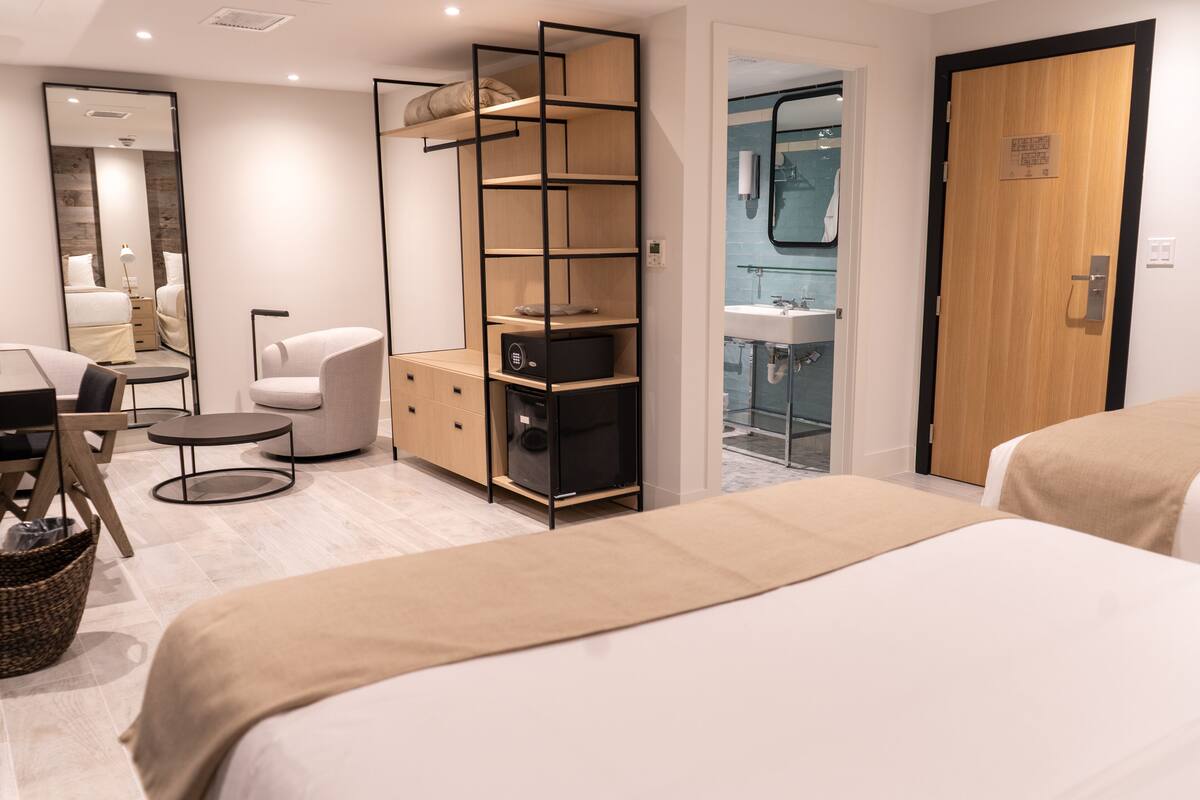 A modern hotel room with a large bed, a desk with a chair, a small round table, a sofa, a tall shelving unit, and a bathroom visible through an open door.