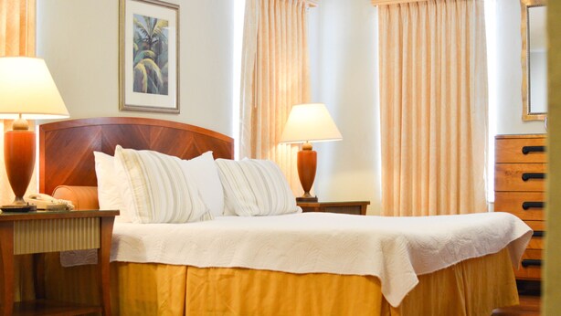 A neatly made bed with a white bedspread and two pillows, flanked by two bedside tables with lamps, a framed picture on the wall, and a wooden dresser. - Viscay Hotel (Miami Beach)