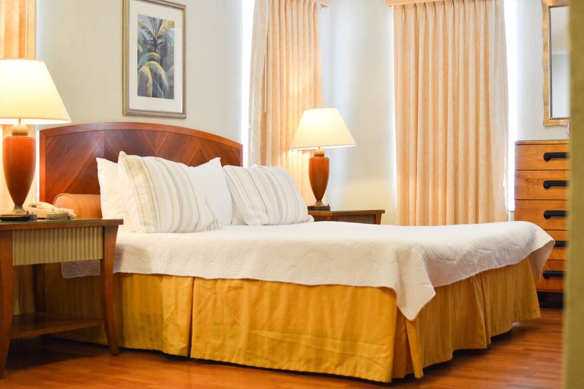 A neatly made bed with a white bedspread and two pillows, flanked by two bedside tables with lamps, a framed picture on the wall, and a wooden dresser.