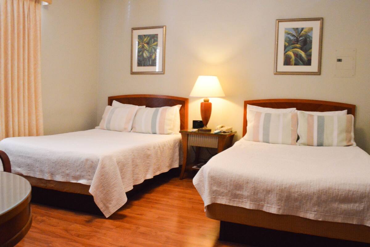 A hotel room with two single beds, a wooden headboard, a nightstand with a lamp, and two framed pictures on the wall.
