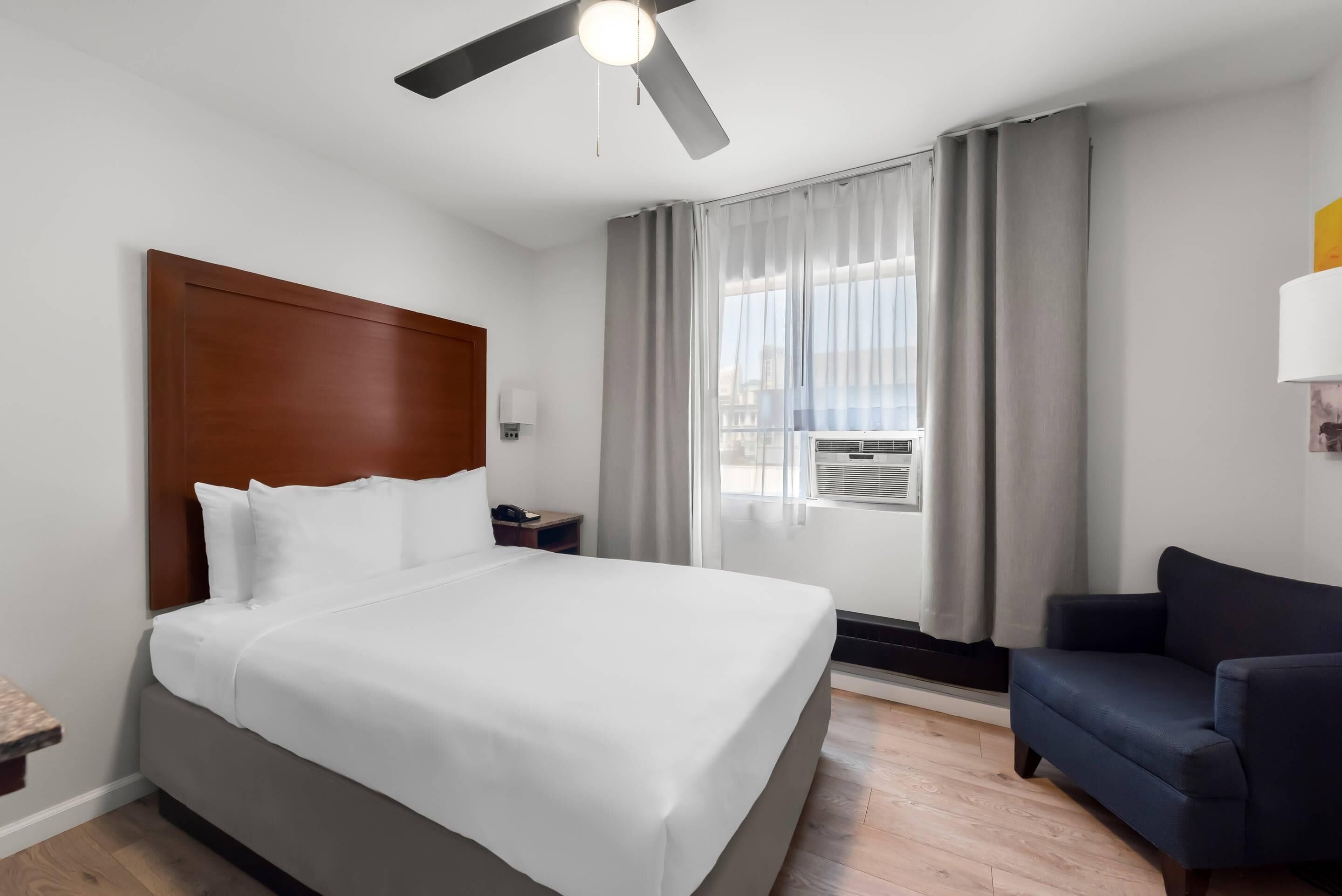 A hotel room with a large bed, a ceiling fan, a window with curtains, a small table, and a dark-colored sofa.