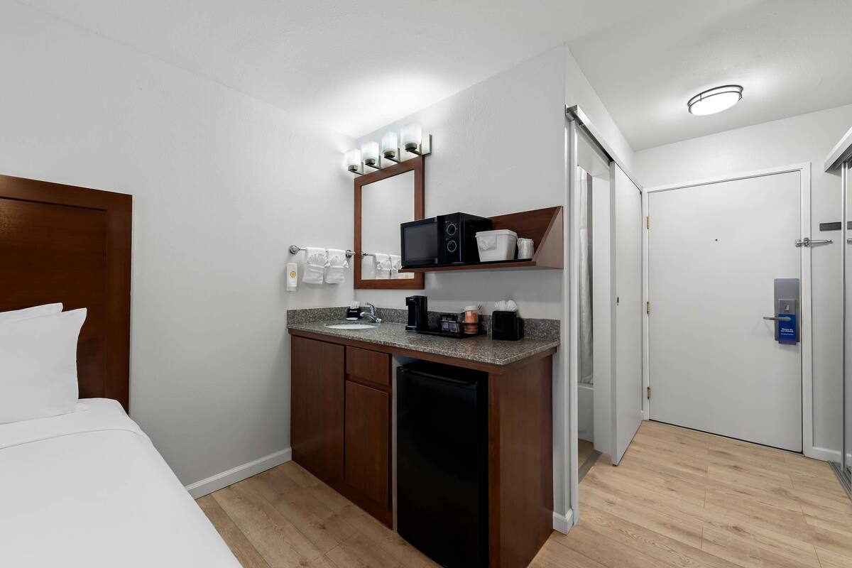 A hotel room with a bed, a small kitchenette with a microwave, a coffee maker, and a mini-fridge. There is a mirror above the counter and a door leading to another room.
