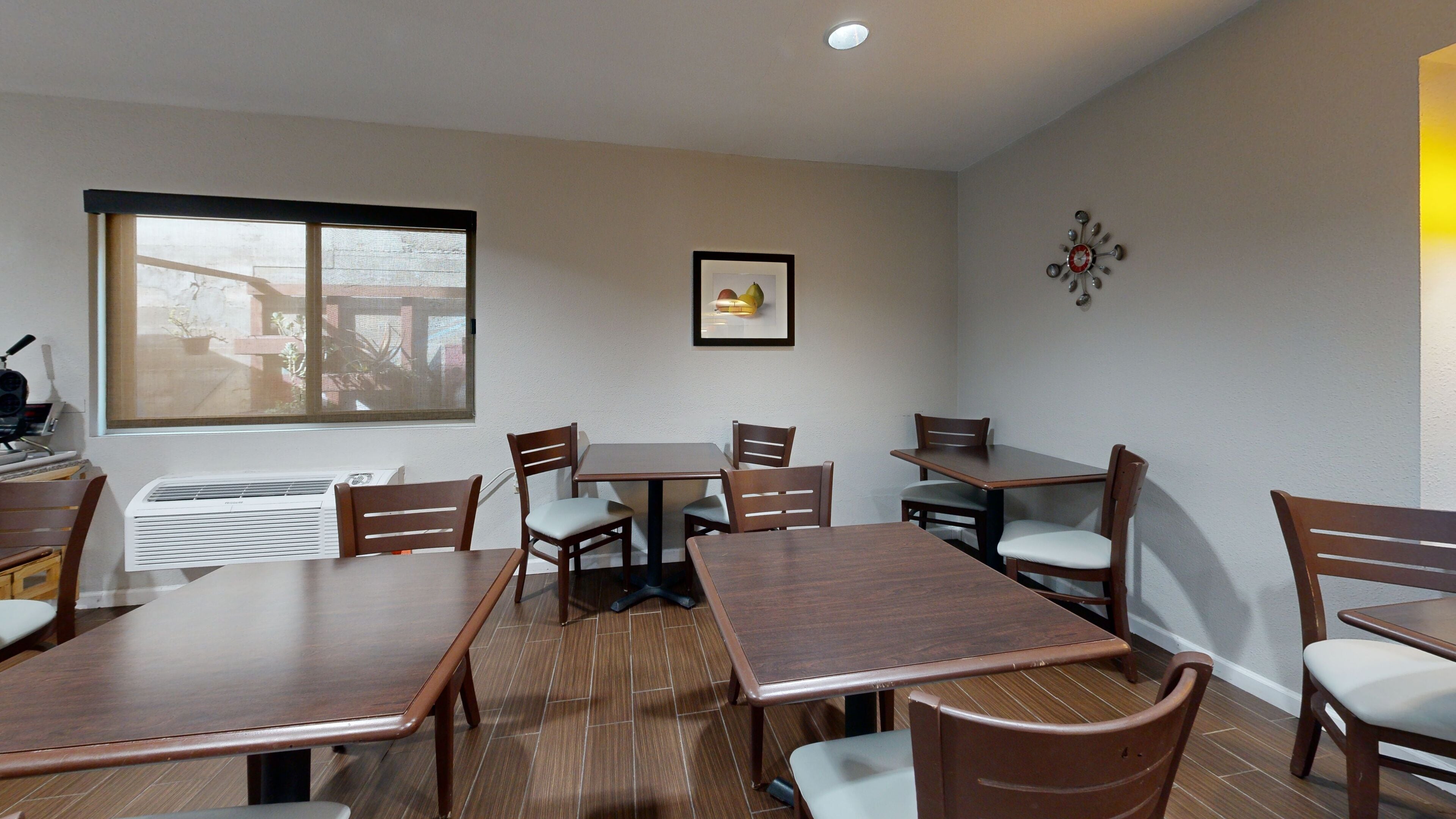 A dining area with wooden tables and chairs, a framed picture on the wall, and a window with a view of buildings.