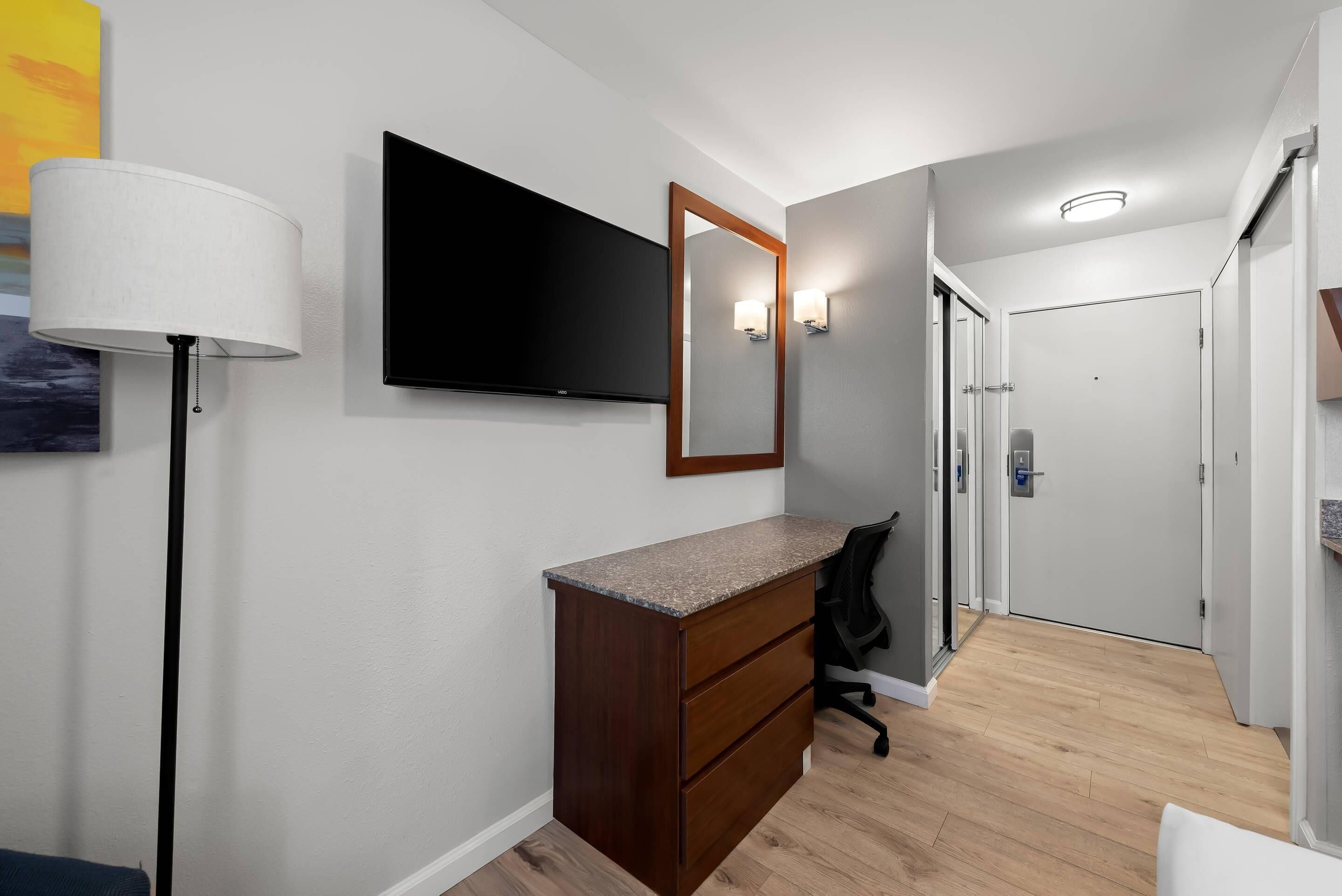 A modern hotel room with a flat-screen TV, a large mirror, a desk with drawers, a chair, and a lamp.