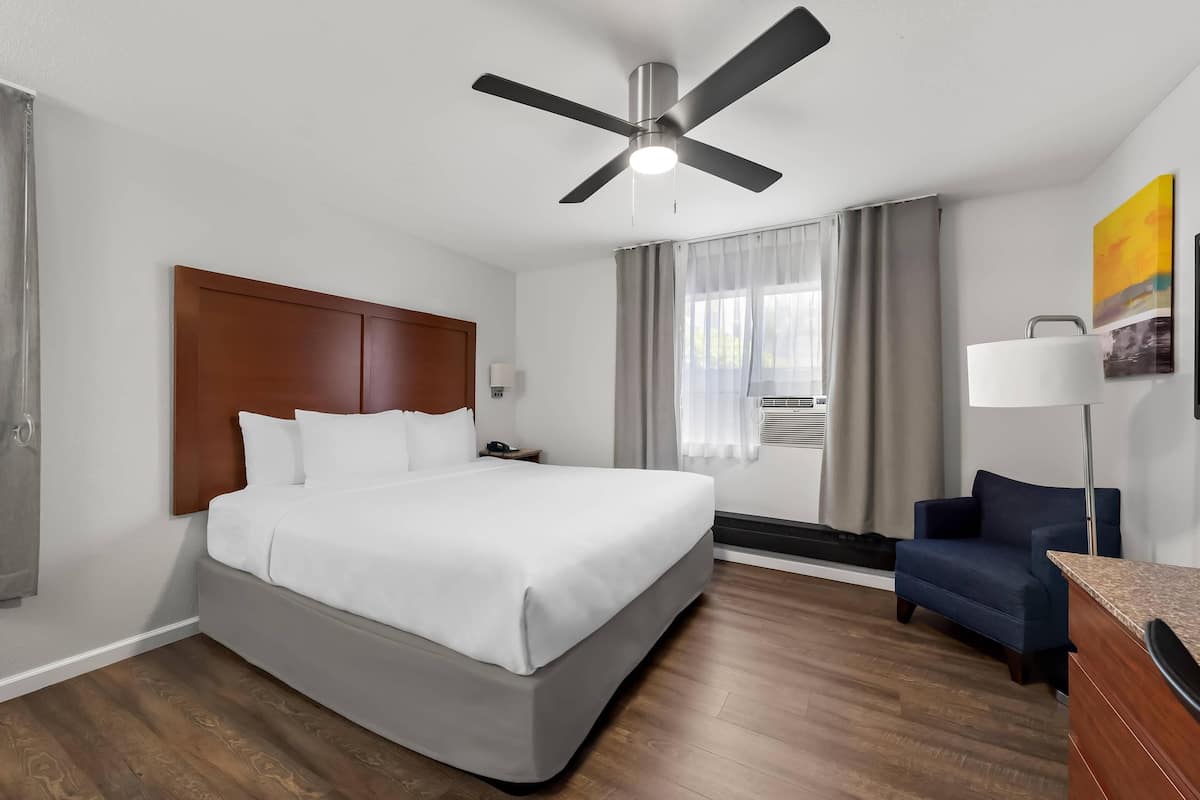 A hotel room with a large bed, a ceiling fan, a desk with a lamp, a chair, and a painting on the wall.