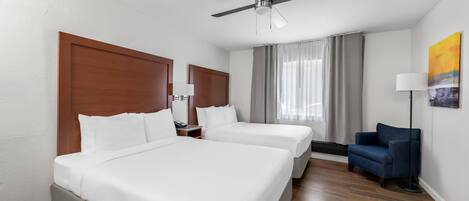 A hotel room with two beds, a ceiling fan, a blue armchair, and a window with curtains.