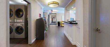 A hotel corridor with a laundry room on the left, a reception desk with a 'Welcome' sign on the right, and a staircase leading to an upper floor.