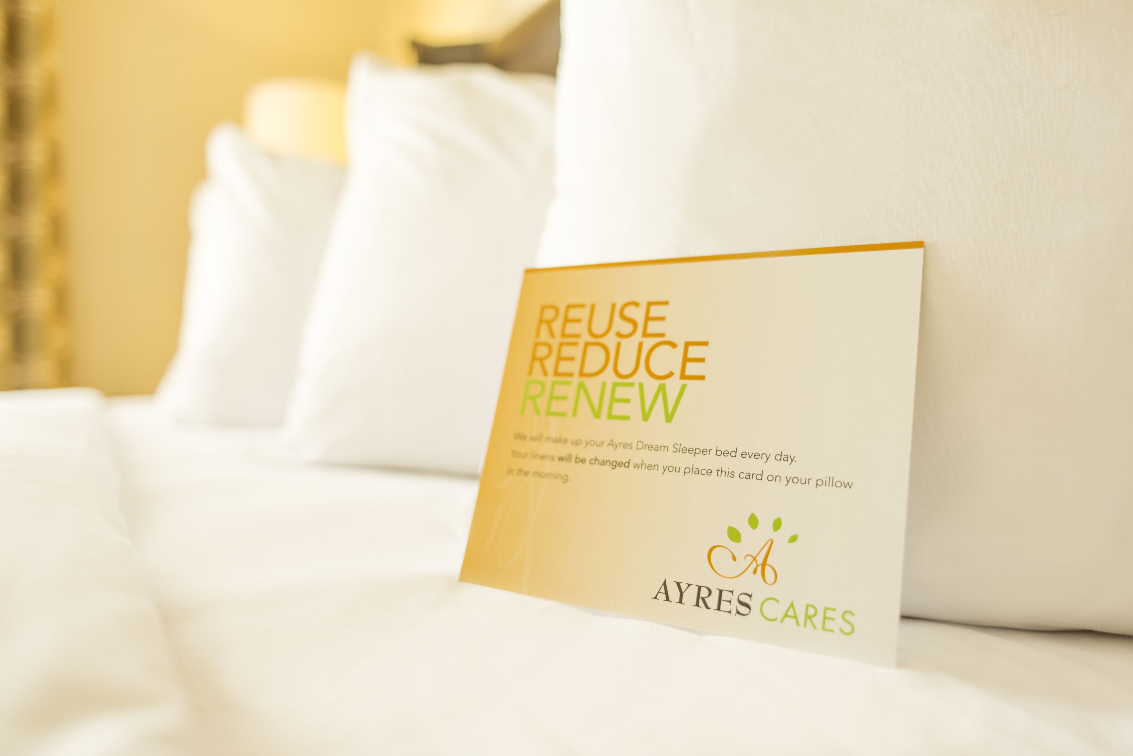 A card on a bed with the text "REUSE REDUCE RENEW" and the logo "AYRES CARES".